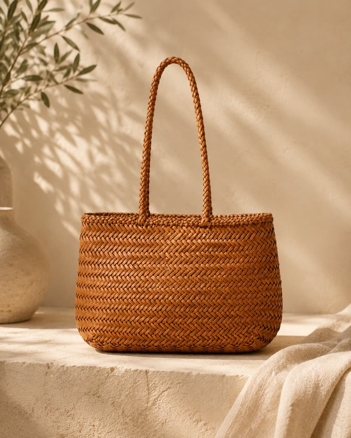 The Capri caramel hand-woven leather basket tote on a sunlit stone ledge, cast with olive-branch shadows